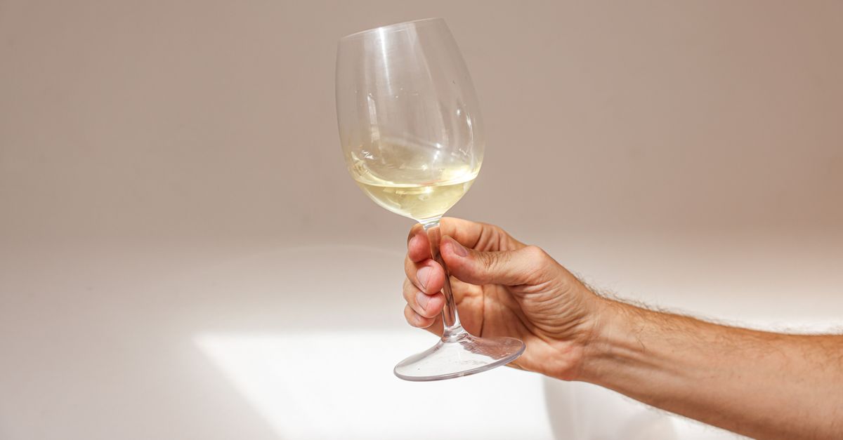 A hand holds a clear wine glass with white wine up toward soft window light against a neutral background.