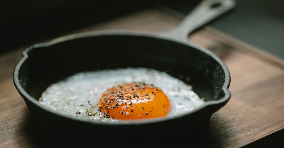 A dark cast iron skillet with a matte, well-seasoned finish holding a frying egg with a bright orange yolk, resting on a wooden surface.