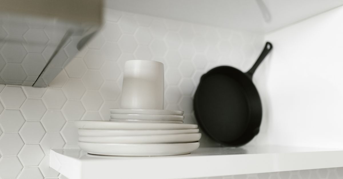 A black cast iron skillet stored on an open white shelf beside a stack of plates against hexagonal tile.