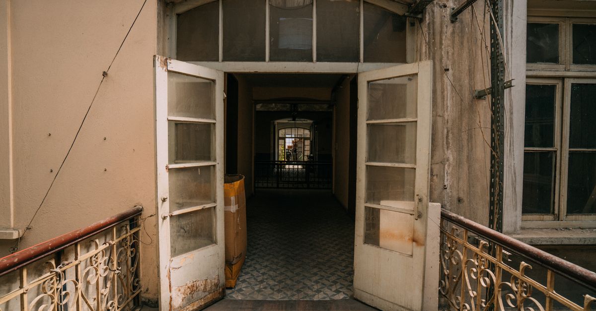 A weathered pair of open glass-paneled wooden doors with peeling paint at the frame, leading into a dim, worn hallway.