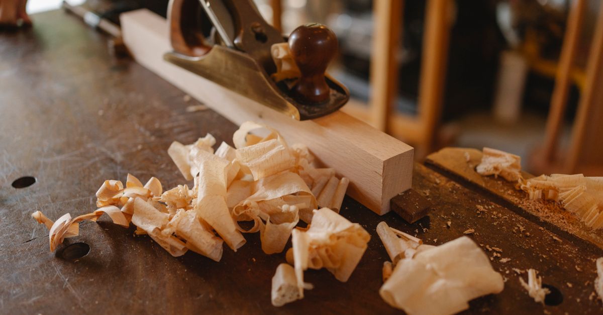 A hand plane rests on a wooden strip on a workbench, surrounded by curled fresh wood shavings.