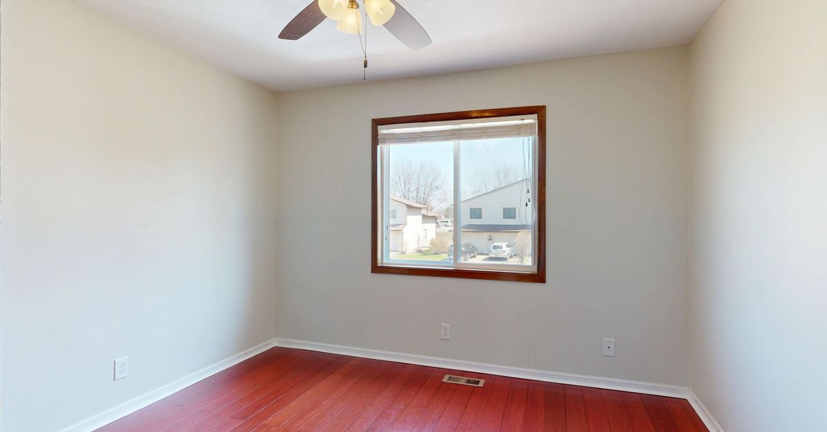 A ceiling fan with a light kit mounted on a white ceiling in a bright, mostly empty room with a window and wood flooring.