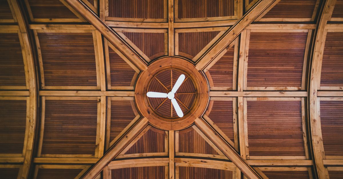 A ceiling fan with white blades mounted in a wooden paneled ceiling with geometric beams, viewed from below.