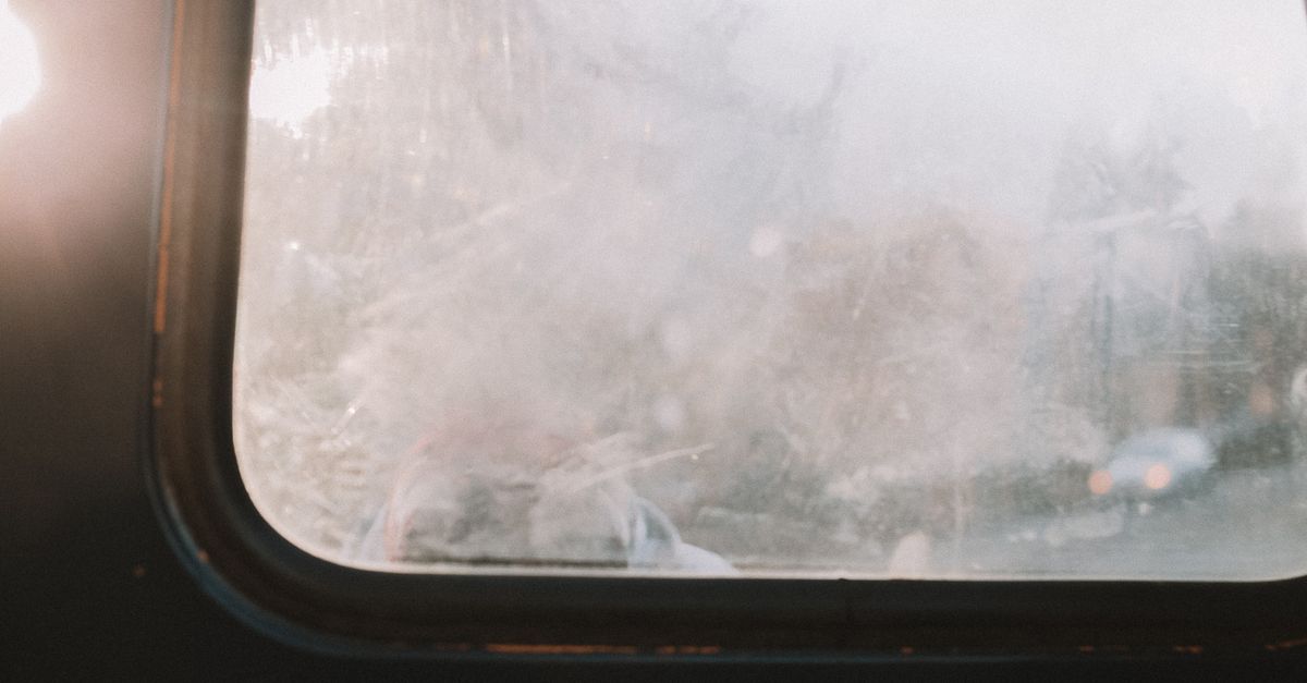 A close-up of a window pane covered in a hazy, grimy film with blurred outdoor scenery visible through the glass.