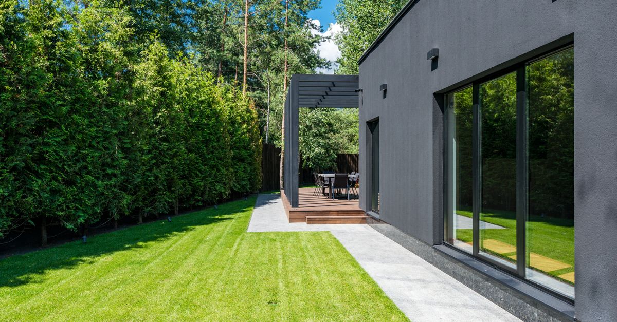 A modern dark gray house with large sliding glass windows reflecting the surrounding green lawn and trees under a clear sky.