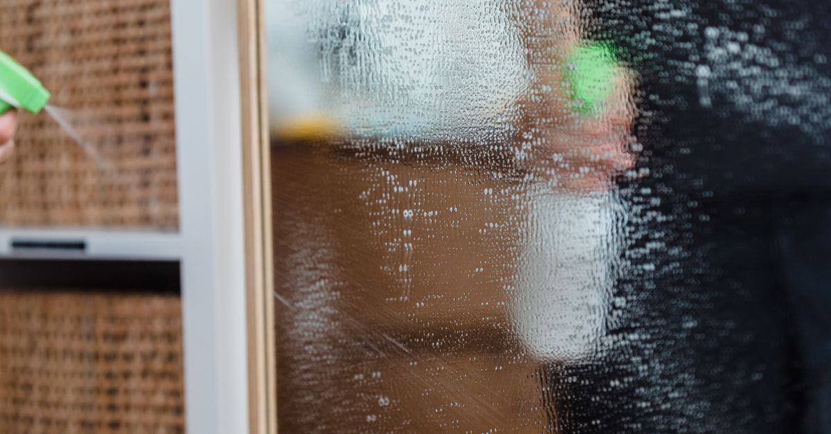A person sprays and wipes a glass window pane with a green spray bottle and cloth, leaving visible cleaning streaks on the glass.