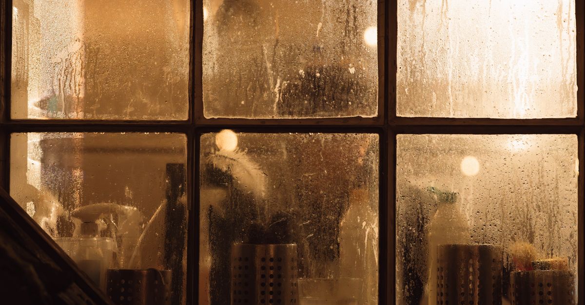 A kitchen window covered in condensation droplets at night, with kitchen items visible blurred through the wet glass.