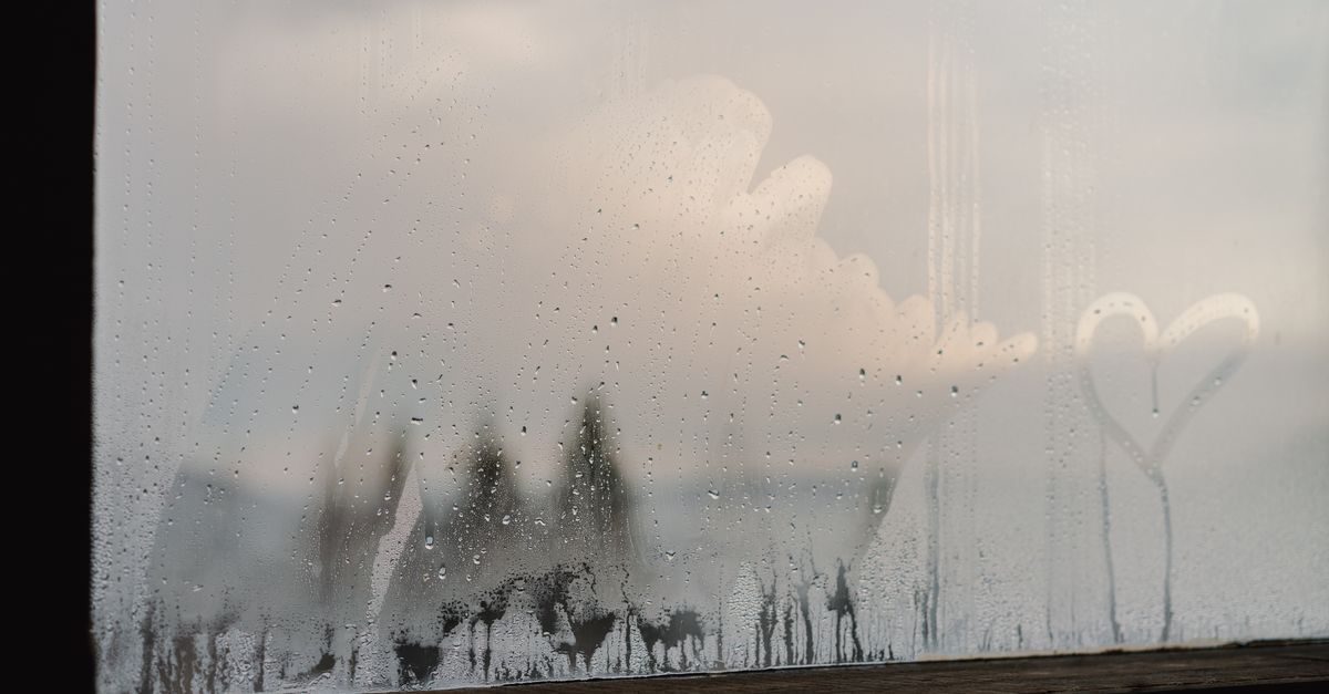 A window pane covered in condensation droplets with a heart shape drawn in the mist, overlooking a blurred outdoor landscape.