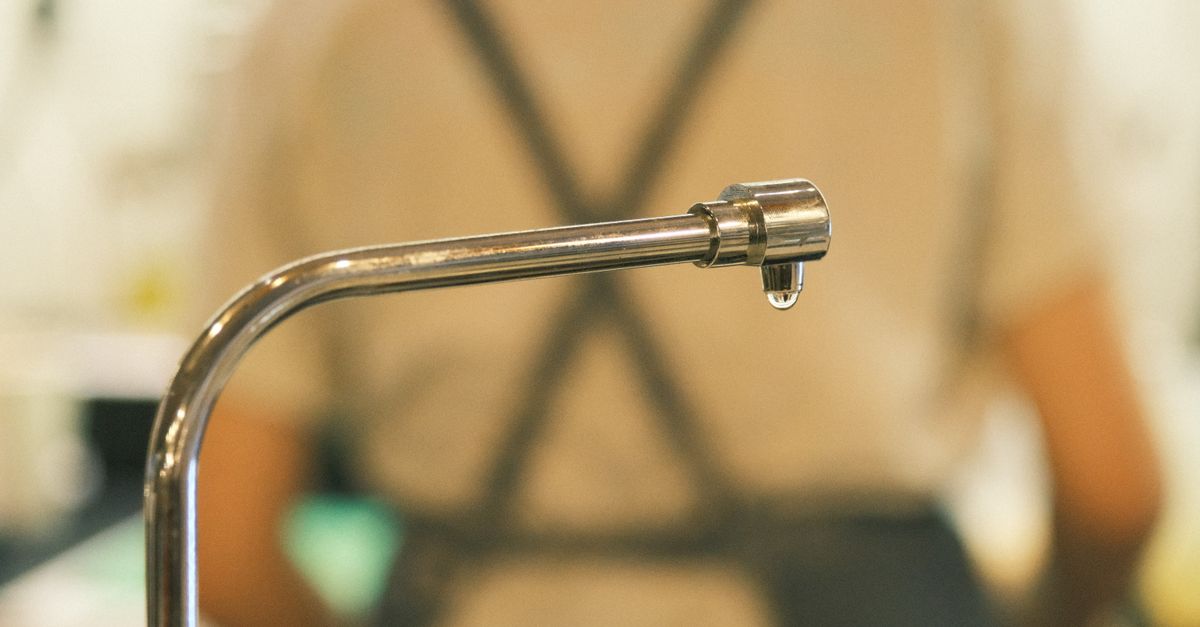 Close-up of a stainless steel kitchen faucet spout with a water droplet forming at its tip, with a blurred person standing behind it.