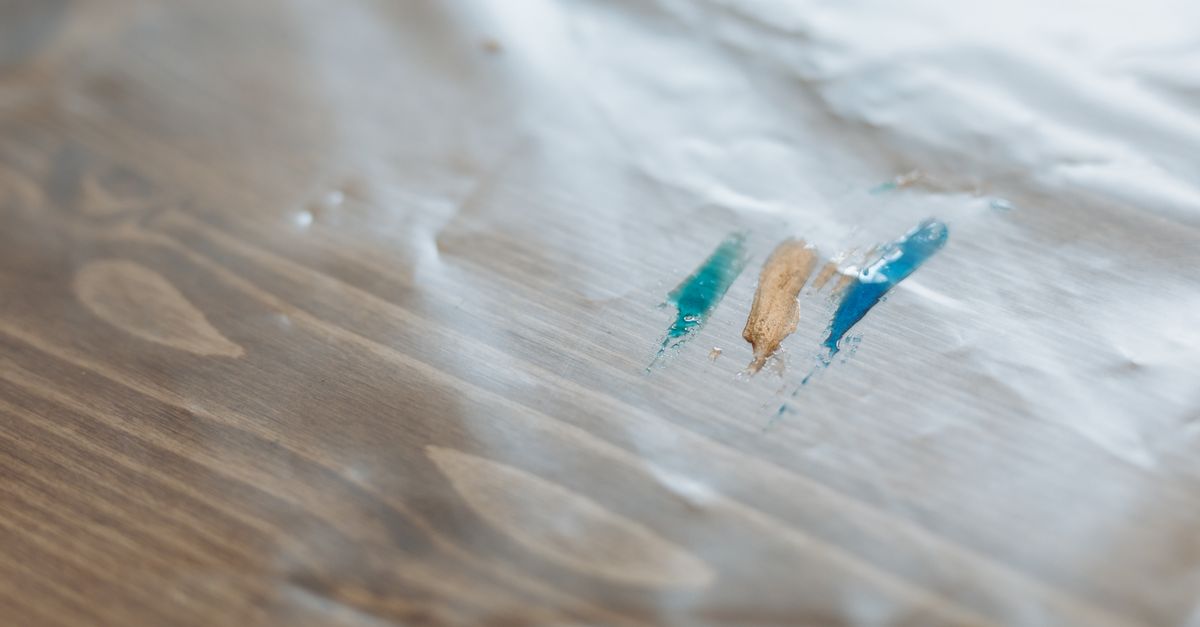 A sheet of clear plastic wrap laid flat on a wooden table surface, with colorful paint strokes visible beneath it.