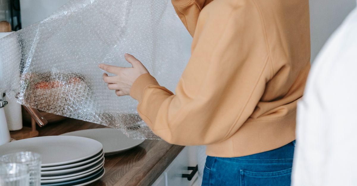A person in an orange sweater lifts a large sheet of clear plastic wrap over a kitchen counter with stacked plates nearby.