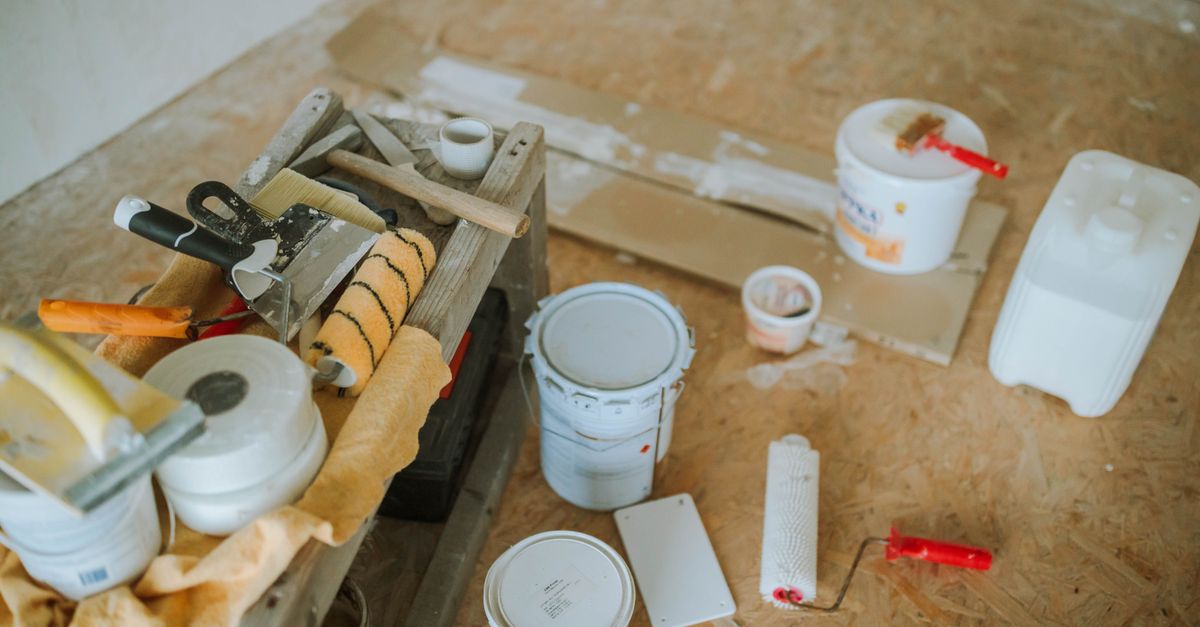 A workbench with assorted painting and drywall supplies including paint buckets, rollers, tape, putty knives and a spreading tool.
