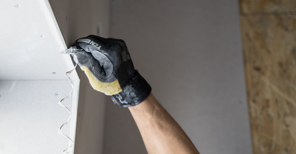 A gloved hand uses a tool to pry a piece of drywall away from a wall framed with wood studs.