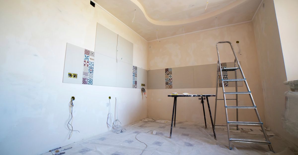 A room mid-renovation with plastered walls, exposed wiring, decorative tile panels, a ladder, and a work table covered in drop cloths.