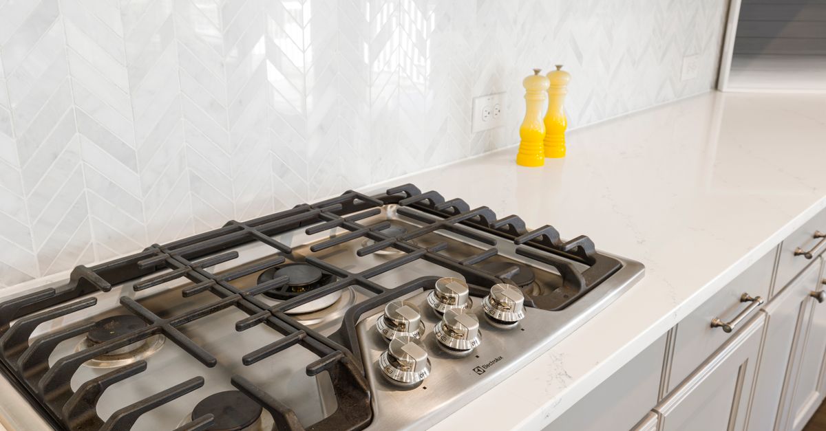 A gas stovetop with cast-iron grates sits against a white chevron-patterned tile backsplash, with two yellow grinders nearby on the countertop.