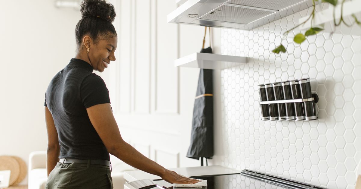 A woman wipes a black induction stovetop with a cloth in a modern white-tiled kitchen.