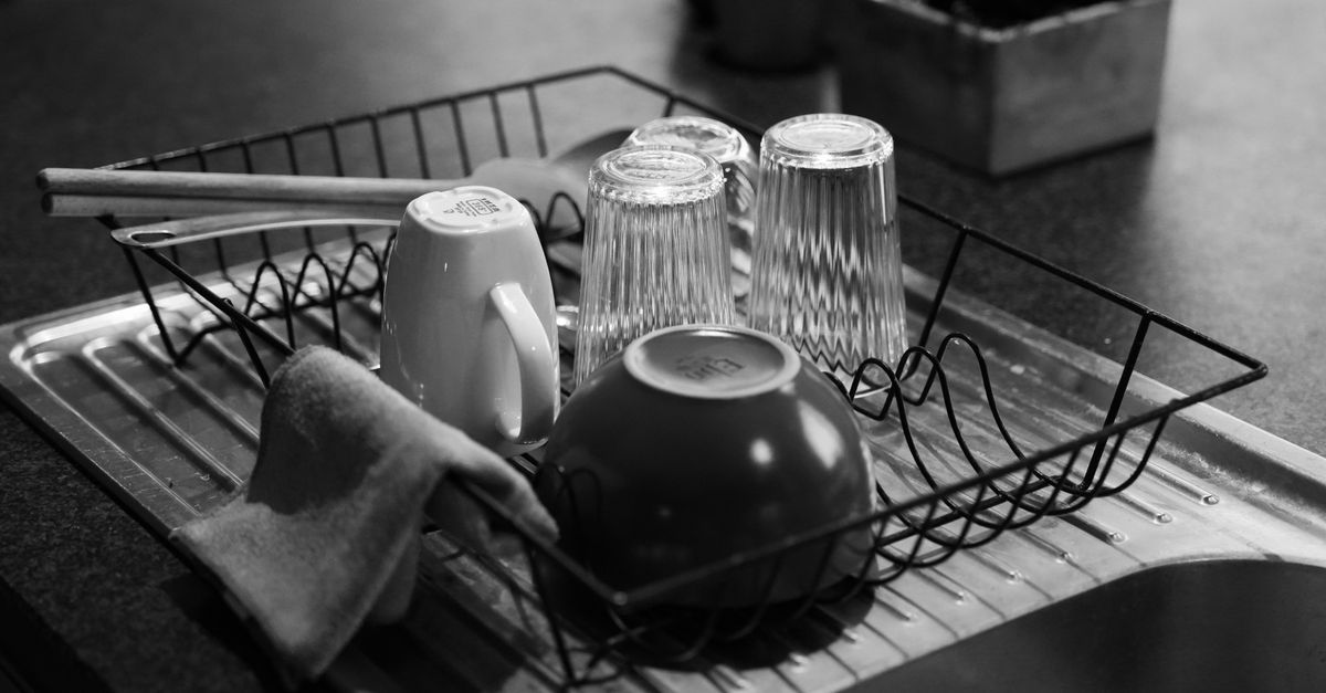 A black-and-white photo of glasses, a mug, and a bowl drying upside down in a metal dish rack beside a folded kitchen towel in a sink.